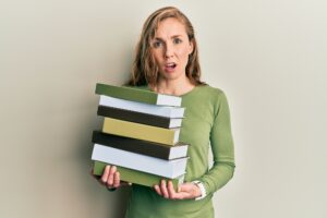 Young blonde woman holding a pile of books in shock face, looking skeptical and sarcastic, surprised with open mouth