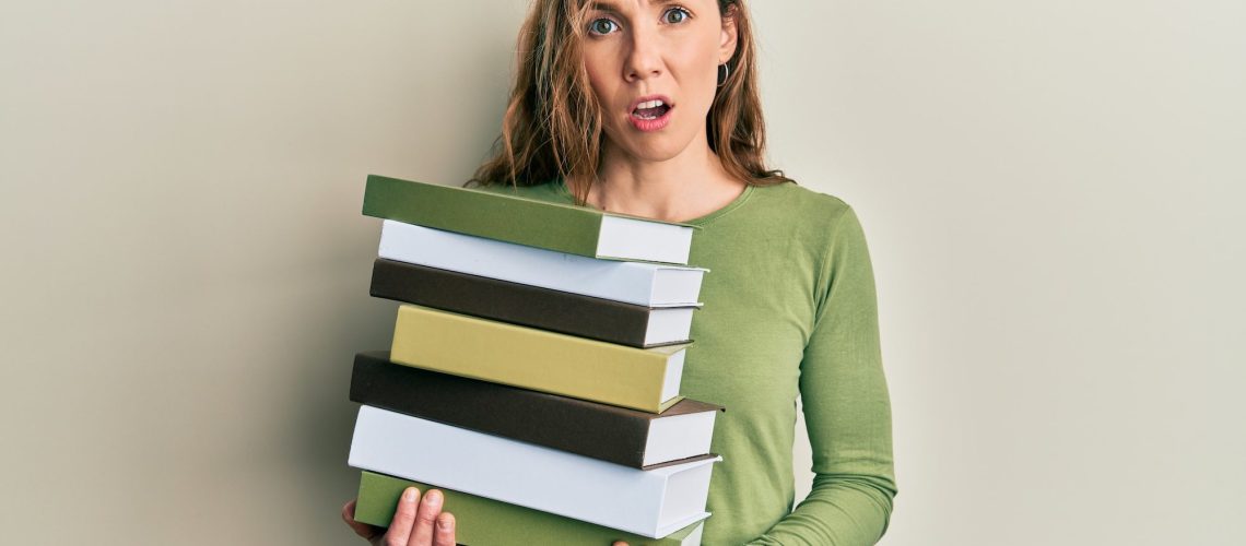 Young blonde woman holding a pile of books in shock face, looking skeptical and sarcastic, surprised with open mouth